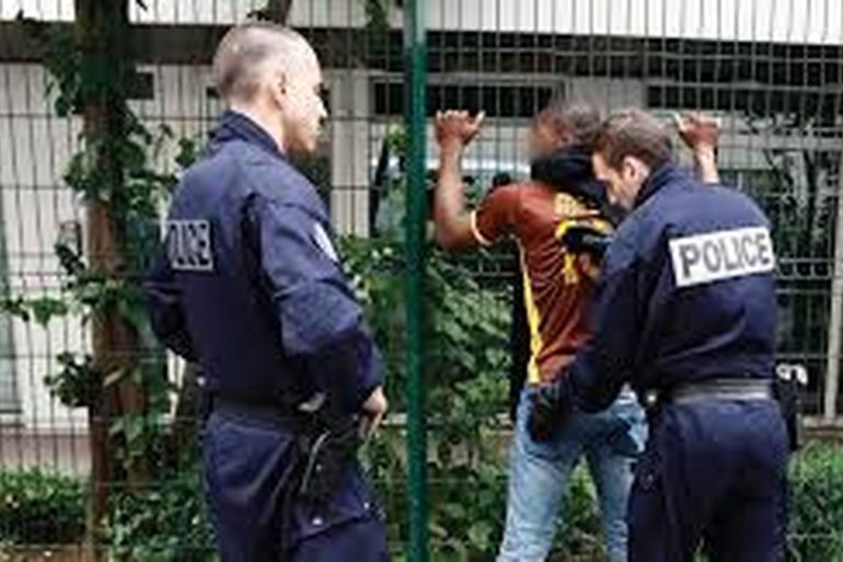 Deux policiers procèdent à un contrôle d’identité sur la voie publique, un citoyen présente ses papiers pendant qu’un autre observe calmement la scène.